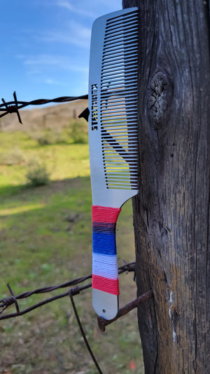 A stainless steel comb with 'STEELTOOTH' etched at the top, suspended on a weathered wooden post by red, white, and blue hemp cord wrapped around the handle. The comb is set against a soft-focus background of a peaceful countryside scene.