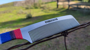Close-up of a stainless steel comb with 'STEELTOOTH' etched on it, handle wrapped in red, white, and blue hemp cordage, resting on a rustic wire against a blurred natural background.