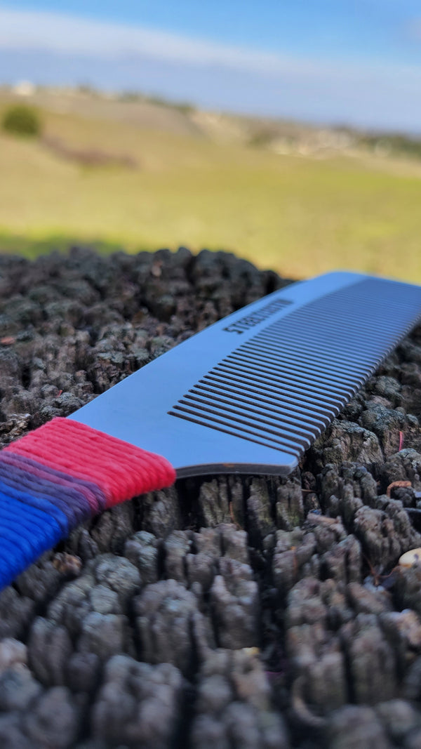 A sleek stainless steel comb lies atop a rugged, textured surface, with a red, white, and blue hemp cord wrapped around the handle. The comb is sharply in focus against a blurred natural landscape in the background, embodying a blend of ruggedness and patriotic elegance.